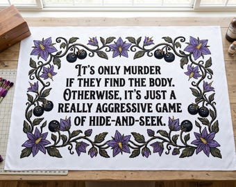 May include: A white fabric panel featuring a black embroidered quote that reads: It's only murder if they find the body. Otherwise, it's just a really aggressive game of hide-and-seek. The text is framed by a border of purple flowers and dark berries.