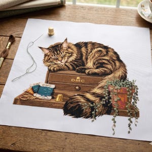 Könnte beinhalten: Kreuzstich eines schlafenden Tabby-Katze auf einer Holzkiste mit der Aufschrift "DMC". Der Kreuzstich zeigt auch eine kleine Topfpflanze, Garnrollen, einen Fingerhut und eine Nadel mit Faden. Der Hintergrund ist weißer Stoff.