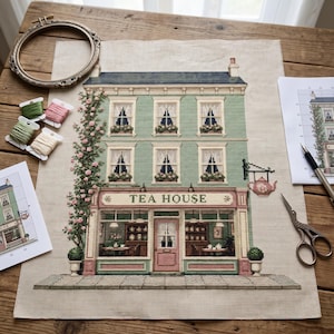 May include: A cross-stitch embroidery project on fabric depicting a three-story green tea house building with a rose vine, a hanging teapot sign, and the text TEA HOUSE. Includes an embroidery hoop, thread bobbins, scissors, and pattern charts.