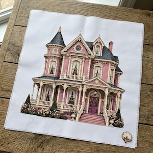 以下が含まれることがあります： 白い布に施されたクロスステッチ刺繍で、濃い青色の屋根、ラップアラウンドポーチ、白い手すり、詳細な窓を持つ多層階のピンクの家が描かれています。右下隅には小さな木製の綿花のアイコンが取り付けられています。