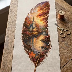 May include: A rectangular piece of beige Aida fabric featuring a cross-stitch design of a large feather. Inside the feather is a landscape with a church, trees with autumn foliage, and a river. Gold-toned scissors and a spool of thread are nearby.