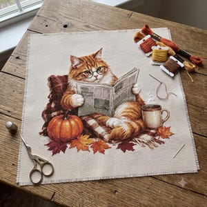 Può includere: Un progetto di ricamo a punto croce su tessuto beige raffigurante un gatto tigrato arancione con occhiali che legge un giornale. La scena include una zucca, foglie autunnali, una tazza, filati da ricamo, un ago e forbici in metallo.