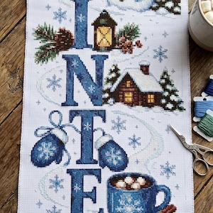 May include: A cross-stitch embroidery piece on white fabric featuring the word WINTER in large blue letters. Each letter incorporates seasonal motifs: a snowman, a lantern, a cabin, blue mittens, a mug of hot cocoa, and white ice skates.
