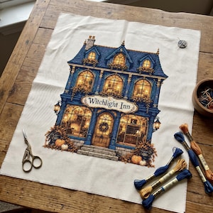 May include: A cross-stitch project on white fabric featuring a blue building labelled Witchlight Inn with glowing windows, pumpkins, and foliage. The scene includes gold embroidery scissors and several skeins of blue and orange embroidery floss.
