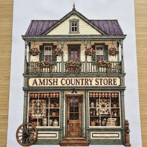 Könnte beinhalten: Eine Kreuzstichstickerei auf weißem Aida-Stoff, die ein zweistöckiges Gebäude mit der Aufschrift AMISH COUNTRY STORE zeigt. Das Bauwerk hat ein violettes Dach, einen grünen Balkon mit Blumenampeln, große Schaufenster und ein hölzernes Wagenrad.