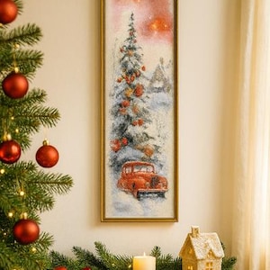 May include: A framed winter scene painting featuring a red vintage car, a snow-covered evergreen tree, and a small house. The painting is displayed above a wooden table with a lit candle, a decorative house, and a wrapped gift.