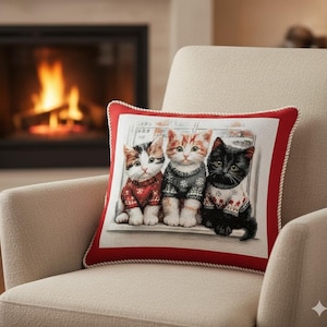 May include: A cushion with a print of three kittens in sweaters. The cats are a calico, ginger tabby, and black cat. The pillow has a red border with a white rope trim. It's on a beige armchair, with a fireplace in the background.