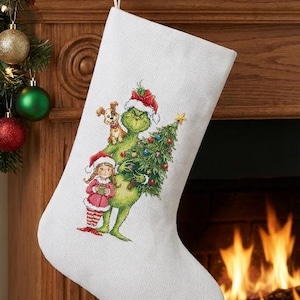 May include: White Christmas stocking with a festive design featuring the Grinch, a girl, and a dog. The Grinch is holding a Christmas tree. The stocking is hanging above a fireplace with a fire burning inside.