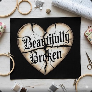 May include: Black cross-stitch fabric with a cracked heart design. The heart is beige with the words "Beautifully Broken" in black gothic lettering. Embroidery hoops, thread, and other craft supplies surround the fabric.