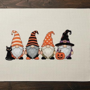 Peut inclure: Un motif de point de croix avec quatre gnomes d'Halloween portant des chapeaux orange, noirs et blancs, un chat noir et une citrouille. Le motif est sur un tissu beige, avec du fil à broder et des ciseaux à proximité.