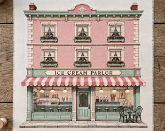 May include: A cross-stitch embroidery pattern on white fabric depicting a three-story building with a pink facade, green trim, and a striped awning. The ground floor features a storefront with the text ICE CREAM PARLOR and interior details.