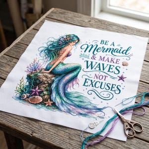 May include: A white fabric square featuring a cross-stitch design of a mermaid with teal hair sitting on a rock, surrounded by coral and seashells. The text reads BE A Mermaid & MAKE WAVES NOT EXCUSES. Includes loose teal and purple thread.