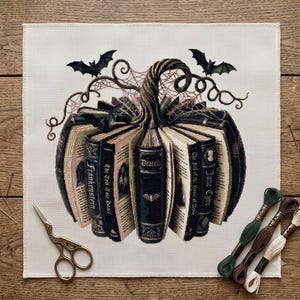 May include: A cross-stitch embroidery on white fabric depicting a pumpkin shape formed by open books with titles like Dracula, Frankenstein, and Jane Eyre. The design includes black bats, spiderwebs, and a stem, accompanied by embroidery scissors and thread.