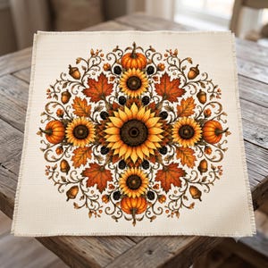 May include: A square piece of off-white Aida fabric featuring a detailed cross-stitch pattern of a circular autumn mandala. The design includes vibrant yellow sunflowers, orange pumpkins, red maple leaves, brown acorns, and small black berries.