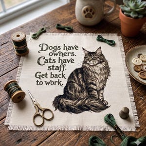 May include: A square piece of beige fabric featuring a cross-stitch design of a long-haired cat and the black text: Dogs have owners. Cats have staff. Get back to work. The fabric has frayed edges and is surrounded by sewing supplies.