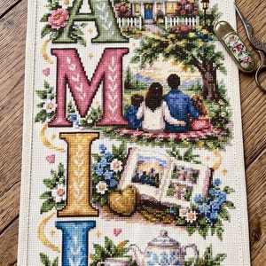Peut inclure: Un échantillon de point de croix vertical sur tissu crème épelant le mot FAMILY en grandes lettres colorées et à motifs. Chaque lettre intègre des scènes détaillées d'une maison, d'une famille sous un arbre, d'un livre et d'un service à thé.