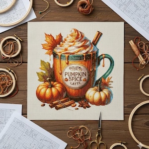 Peut inclure: Motif de point de croix représentant un latte au pumpkin spice dans une tasse en bois, avec de la crème fouettée, une bâton de cannelle et le texte "Pumpkin Spice Latte". Deux citrouilles, des feuilles d'automne et des bâtons de cannelle.