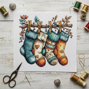 Può includere: Un disegno a punto croce con quattro calze natalizie appese a un ramo di legno con mollette. Le calze sono nei toni del blu, arancione e crema, con vari motivi e dettagli a cuore. L'immagine è circondata da materiali di cucito.