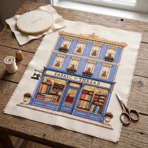 May include: A cross-stitch embroidery piece depicting a blue three-story building with the text FABRIC THREAD on the storefront. The design includes shelves filled with colorful fabric bolts, windows with flower boxes, and a sewing machine sign.