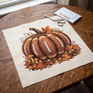 May include: A square piece of cream-colored fabric featuring a cross-stitch design of a pumpkin with football-textured segments, surrounded by autumn leaves and small mushrooms, accompanied by a pair of silver scissors and a color pattern key.