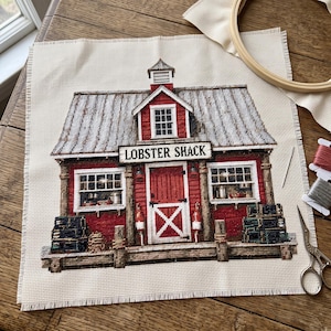 May include: A cross-stitch project on beige fabric depicting a red lobster shack with a gray roof, white trim, and the text LOBSTER SHACK. The scene includes windows, a central door, and stacks of dark crates. Embroidery supplies are visible nearby.