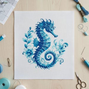 Puede incluir: Patrón de punto de cruz de un caballito de mar en tonos azules, rodeado de elementos florales y de hojas. El caballito de mar está sobre tela blanca, con hilo de bordar, tijeras y agujas. El diseño es detallado e intrincado.