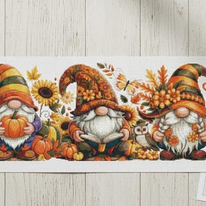 Può includere: Un modello a punto croce con tre gnomi autunnali con zucche, girasoli e foglie autunnali. Gli gnomi indossano cappelli colorati e tengono in mano un cartello con la scritta "Hello Fall". Sono presenti anche telai da ricamo, filo e una tazza di caffè.