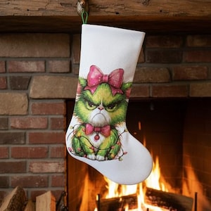 May include: White Christmas stocking featuring a grumpy green character with a pink bow and bow tie, surrounded by string lights. The stocking hangs from a wooden mantel with a deer head hook, in front of a brick fireplace.