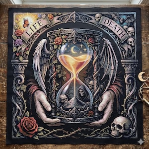 May include: A square cross-stitch embroidery on black fabric featuring a central hourglass held by two hands. The design includes a white angel wing, a dark bat wing, roses, skulls, and banners with the text LIFE and DEATH. The hourglass contains glowing sand, a crescent moon, and stars.