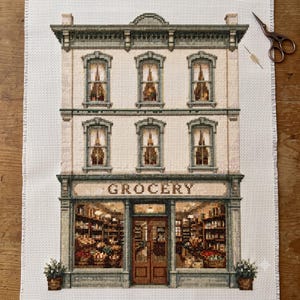 Könnte beinhalten: Eine Kreuzstichstickerei auf weißem Stoff, die ein dreistöckiges Gebäude mit einem Ladenlokal mit der Aufschrift GROCERY darstellt. Das Design zeigt detaillierte Fenster, einen zentralen Eingang und Regale voller Waren. Eine Metallschere liegt daneben.