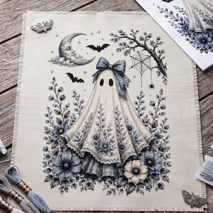 May include: A cross-stitch embroidery project on beige fabric featuring a central ghost figure wearing a large blue bow. The design includes a crescent moon, bats, a spider web, and dark blue floral elements. Embroidery floss and scissors are nearby.