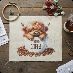 May include: Cross-stitch project featuring a Highland cow calf nestled in a coffee cup, surrounded by coffee beans. The cup reads "I NEED COFFEE." Craft supplies, including embroidery floss, needles, and a wooden hoop, are arranged on a wooden surface.