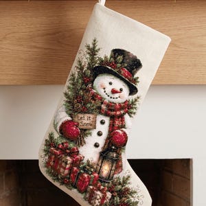 May include: A cream-colored fabric Christmas stocking featuring a detailed cross-stitch design of a snowman wearing a black top hat and red plaid scarf. The snowman holds a sign that reads Let it Snow, a lantern, and is surrounded by gifts.