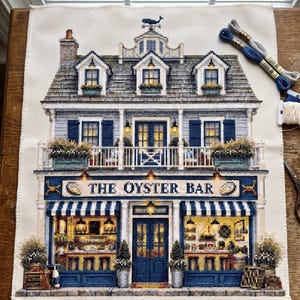 May include: A cross-stitch embroidery project on cream fabric depicting a two-story building labeled THE OYSTER BAR. The design features blue shutters, a balcony with flower boxes, a whale weather vane, and warm yellow interior lighting.