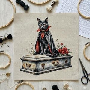 May include: Cross-stitch depicting a black cat in a cape and collar, perched on a coffin. The cat's collar and cape lining are red. Red roses and foliage adorn the coffin's base. Embroidery hoops, thread, and scissors are arranged around the cross-stitch.