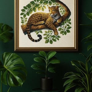 May include: A framed artwork featuring a leopard resting on a tree branch, surrounded by green leaves. The leopard has a tan coat with dark spots. The frame is gold-colored, and the background is a dark green. Potted plants are in the foreground.