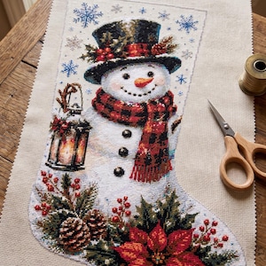 May include: A cross-stitch Christmas stocking on beige fabric featuring a snowman wearing a black top hat and a red and black plaid scarf. The snowman holds a lantern, and the base is decorated with pinecones, holly berries, and a red poinsettia.