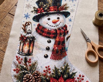 May include: A cross-stitch Christmas stocking on beige fabric featuring a snowman wearing a black top hat and a red and black plaid scarf. The snowman holds a lantern, and the base is decorated with pinecones, holly berries, and a red poinsettia.