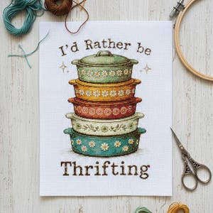 Puede incluir: Arte de punto de cruz que muestra una pila de fuentes de horno vintage con diseños florales. El texto "I'd Rather be Thrifting" está bordado debajo. Hilos de bordar y un bastidor de bordado de madera en el fondo.