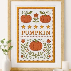 Könnte beinhalten: Gerahmte Kreuzstichkunst mit orangefarbenen Kürbissen, Sternen und floralen Elementen. Das Wort "PUMPKIN" ist in der Mitte zu sehen. Das Kunstwerk ist in einem hellen Holzrahmen gerahmt und ruht auf einem Holzregal.