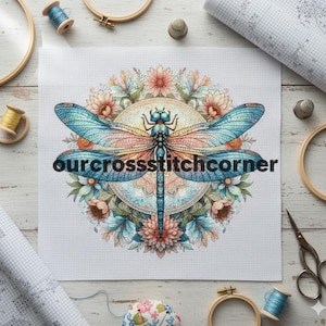 May include: Cross-stitch pattern featuring a dragonfly with blue and orange wings, surrounded by floral elements. The text "ourcrossstitchcorner" is displayed across the design. Crafting supplies, including thread spools, hoops, and scissors, are arranged around the pattern.
