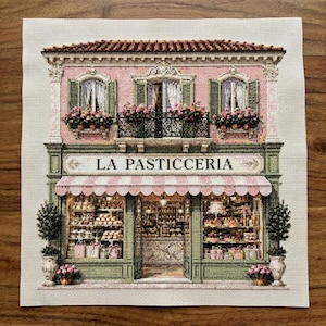 May include: A cross-stitch embroidery of a storefront labelled LA PASTICCERIA, featuring a pink facade, green trim, a striped awning, and windows filled with pastries. The upper level includes a balcony with flower boxes and a tiled roof.