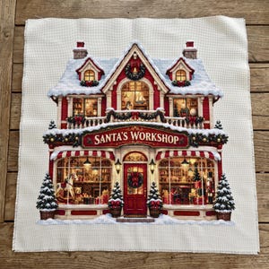 Könnte beinhalten: Ein Kreuzstich-Stoffpanel mit einer detaillierten Illustration eines zweistöckigen Gebäudes mit roter Fassade, schneebedecktem Dach und einem Schild mit der Aufschrift SANTA'S WORKSHOP. Die Szene zeigt dekorierte Fenster, Tannenbäume und Spielzeug.