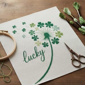 Può includere: Progetto di punto croce con un disegno di dente di leone verde e la parola "lucky". Il disegno include fiori a forma di trifoglio. Filo da ricamo, forbici e un telaio di legno sono anch'essi presenti, suggerendo un progetto artigianale.