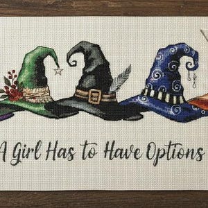 Può includere: Un'opera d'arte ricamata con cinque cappelli da strega colorati in viola, verde, nero, blu e arancione. La frase "A Girl Has to Have Options" è ricamata sotto. Forniture per cucito, tra cui rocchetti di filo e forbici, circondano l'opera.