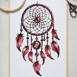 Puede incluir: Punto de cruz de un atrapasueños en burdeos y rosa. El diseño presenta una red central, cuentas y plumas colgantes. La obra de arte está enmarcada en un marco de madera clara. Medidas aproximadas: 30 cm x 40 cm.