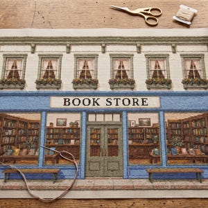 Könnte beinhalten: Eine Stickvorlage, die die Fassade eines Buchladens mit blauer Front, großen Schaufenstern mit Blick auf Bücherregale und dem Schriftzug BOOK STORE zeigt. Das Design umfasst eine Kletterpflanze, zwei Bänke und Fenster im Obergeschoss.