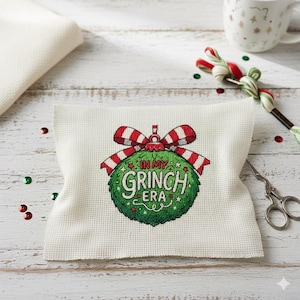 Könnte beinhalten: Ein weißer Kreuzstichstoff mit einem grünen Weihnachtsbaumschmuck-Design mit der Aufschrift "In My Grinch Era". Der Schmuck hat eine rot-weiß gestreifte Schleife. Weitere Artikel sind eine Tasse, Zuckerstangen und eine Schere.