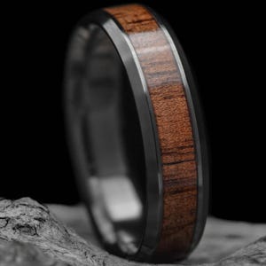 May include: A ring featuring a dark metal band with a central inlay of reddish-brown wood. The wood grain is clearly visible. The ring is presented against a dark backdrop, emphasizing its design and texture.
