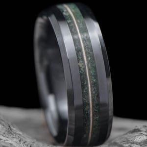 May include: A black ring featuring a central band of green stone and a silver string inlay. The ring has a polished finish, set against a dark background.